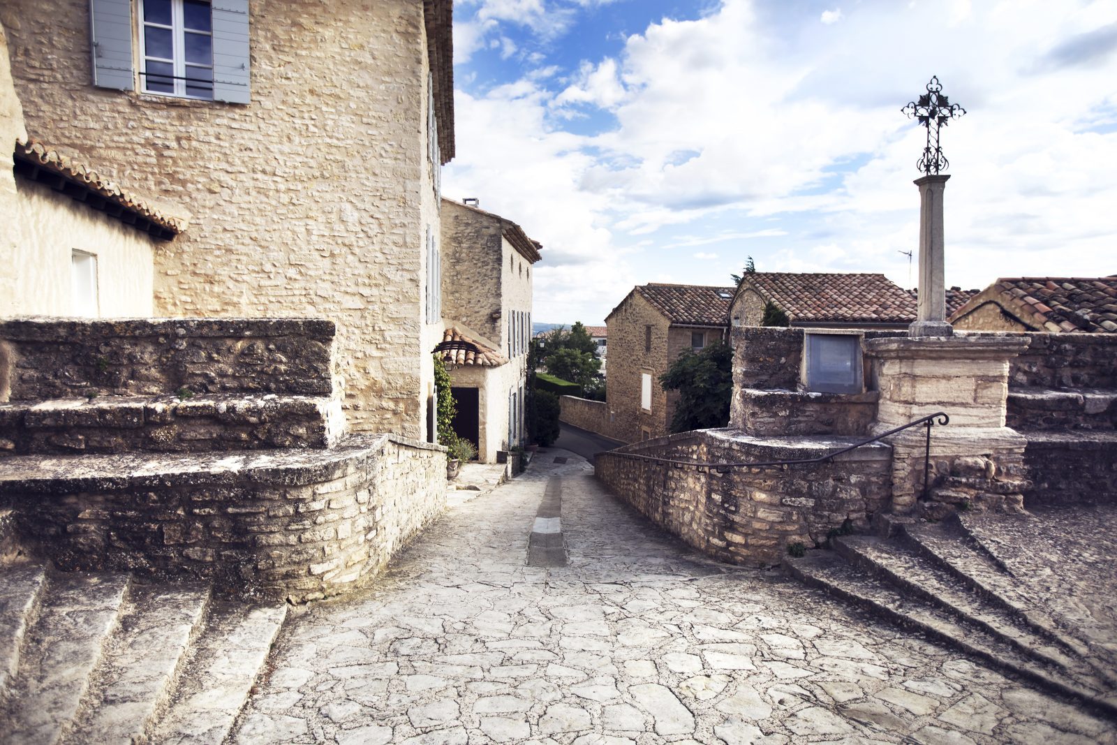 A traditional Provençal cobblestone street in Gordes, with a tall iron cross at the right and stone houses on the left
